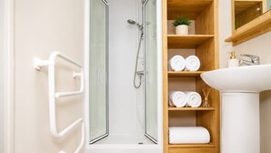 Un bany amb dutxa tancada per vidre, una prestatgeria de fusta amb tovalloles, un lavabo blanc i un mirall sobre el lavabo.