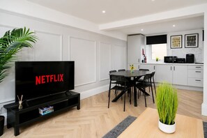 Ett modernt vardagsrum med en platt-TV som visar ”NETFLIX”, en matplats med ett runt bord och stolar samt ett kök med vita skåp och vitvaror.