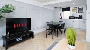 Un saló modern amb televisió de pantalla plana, zona de menjador amb taula i cadires, i cuina amb armaris i electrodomèstics.