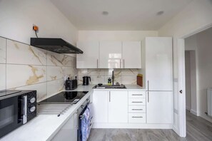 A compact kitchen with white cabinets, a black range hood, a microwave, and a kettle.