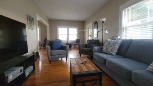 Ein Wohnzimmer mit Fernseher, Sofas, Sesseln, einem Couchtisch und einem Fenster mit Jalousien.
