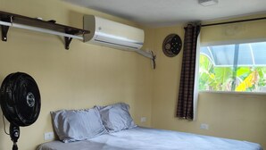 Ein Schlafzimmer mit einem Bett, einem Standventilator, einer Klimaanlage, einem Fenster mit Vorhängen und einer Wanduhr.