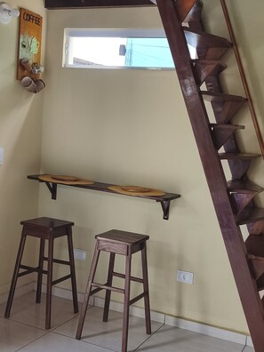 A staircase with a wooden handrail, a window with a view of a building, a wall-mounted shelf with mugs, and two wooden stools. - Cozy Chalet, Your Beach Retreat (Cabo de Santo Agostinho)
