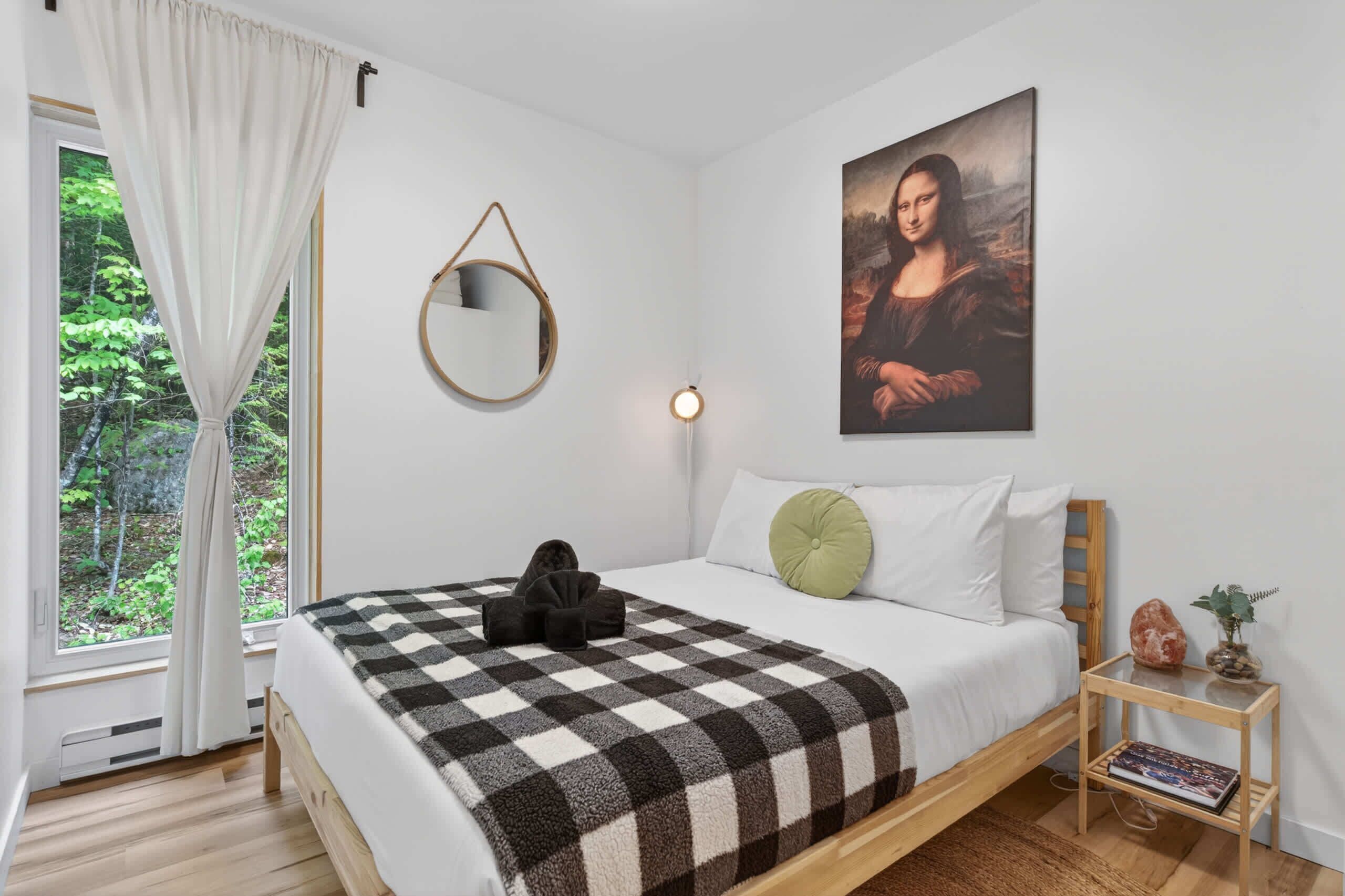 A bedroom with a bed, a painting, a mirror, a bedside table with a book, and a window with curtains.