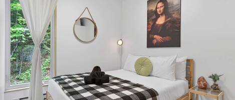 A bedroom with a bed, a painting, a mirror, a bedside table with a book, and a window with curtains.