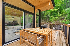 A wooden deck with a table and chairs, large sliding glass doors, and a view of a forest. - Cabin Retreat & Spa | La Cozy Creek Cabin (La Conception)