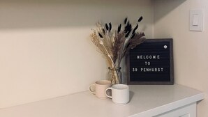 A kitchen with white cabinets, black handles, a welcome sign, a vase with dried flowers, and two mugs.