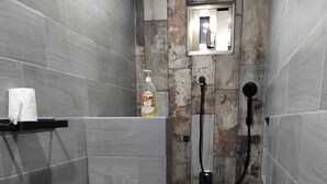 Een smalle douchecel met een toilet aan de linkerkant, een zeepdispenser op de muur en een klein raam boven de spiegel.