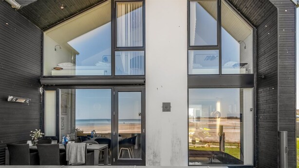 A modern building with large glass windows, a deck area with outdoor furniture, and a view of the beach.
