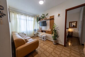 A living room with a sofa, a TV, a small table, and a potted plant. - Apartamento com Garagem a 450m da Praia do Forte (Praia Grande)