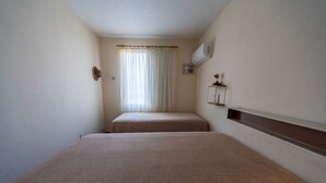 Une chambre d’hôtel étroite, avec un lit simple, une fenêtre avec des rideaux, un climatiseur fixé au mur et une petite étagère avec quelques objets.
