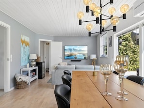 Ein modernes Wohnzimmer mit einem Holzesstisch, schwarzen Stühlen, einem grauen Sofa, einem Fernseher und einem großen Fenster mit Blick auf Grünflächen.