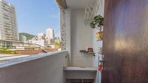 Un balcón estrecho con vistas a edificios de la ciudad, un lavamanos pequeño de color blanco y una planta en maceta.