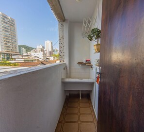 A narrow balcony with a view of city buildings, a small white sink, and a potted plant. - Apartamento com Garagem a 450m da Praia do Forte (Praia Grande)