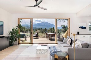 Una sala de estar moderna con un amplio ventanal corredera que da a una terraza exterior, un ventilador de techo y vistas a la montaña.