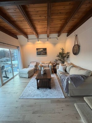 Amplia sala de estar con techo de madera, un ventanal grande, un conjunto de sofás, una mesa de centro y una planta en maceta.