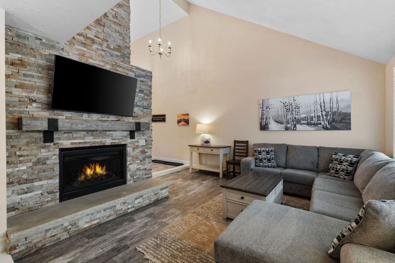 Un salon avec une cheminée en pierre, un téléviseur à écran plat fixé au-dessus, un canapé gris, une petite table et un tableau accroché au mur.