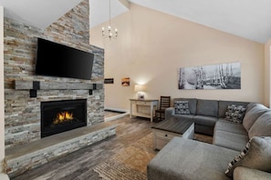 Una sala de estar con chimenea de piedra, un televisor de pantalla plana montado encima, un sofá gris, una mesita y un cuadro en la pared.