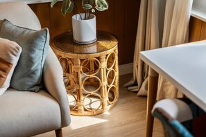 A wicker side table with a potted plant, a white chair, and a white table in a room with wooden flooring and curtains.