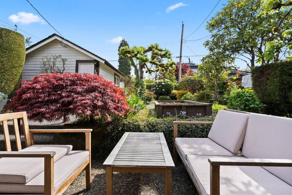 A patio with a wooden table and two sofas, surrounded by various plants and a house.