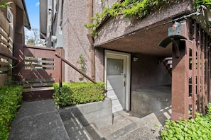 A narrow stairway leading to a door, with a small garden area and a building facade.