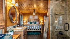 A log cabin interior with a wooden ceiling, a bed with a headboard, a bathroom with a mirror, and a shower area. - Sapphire Sky | Kids Fun Zone | Sauna | Mtn Views (Mineral Bluff)