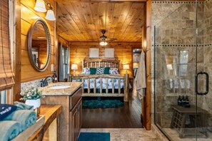 A log cabin interior with a wooden ceiling, a bed with a headboard, a bathroom with a mirror, and a shower area. - Sapphire Sky | Kids Fun Zone | Sauna | Mtn Views (Mineral Bluff)