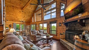 A log cabin interior with a fireplace, wooden furniture, and large windows. - Sapphire Sky | Kids Fun Zone | Sauna | Mtn Views (Mineral Bluff)