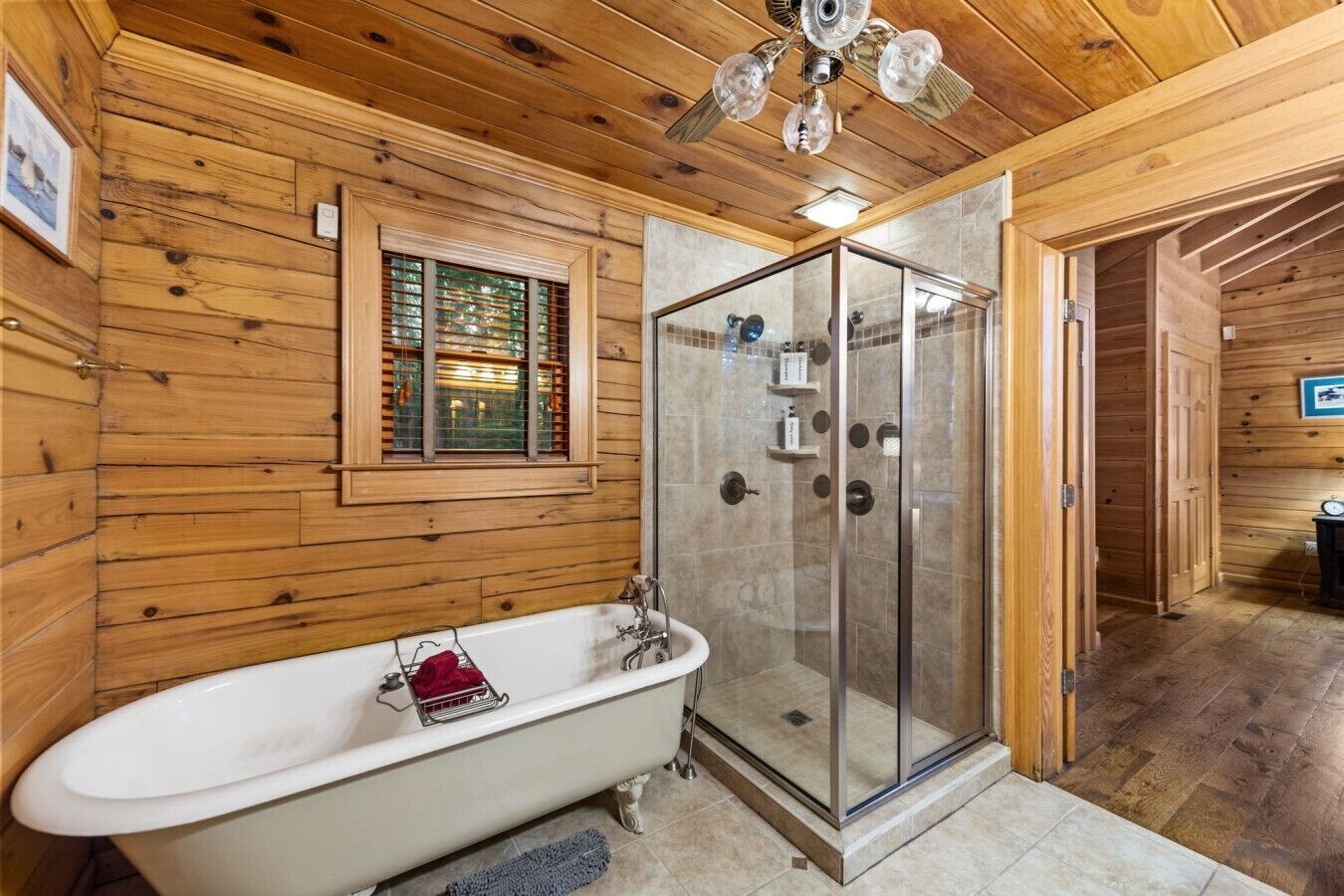 A wooden-paneled bathroom with a freestanding bathtub, a glass-enclosed shower, and a window with blinds.