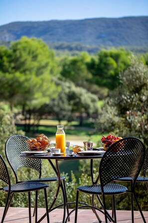 Ein Tisch im Freien mit Frühstück, darunter Croissants, Obst und Saft, mit Stühlen und einer schönen Aussicht im Hintergrund.