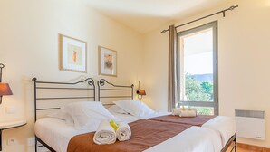 A neatly made bed with white linens and a brown blanket, two framed pictures on the wall, a bedside lamp, and a window with a view of greenery. - Provence Country Club by Interhome (L'isle sur la Sorgue)