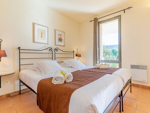 Ein ordentlich bezogenes Bett mit weißen Laken und einer braunen Decke, zwei gerahmte Bilder an der Wand, eine Nachttischlampe und ein Fenster mit Blick ins Grüne.