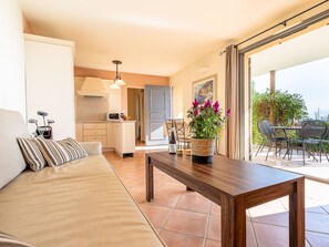 Ein gemütlicher Wohnbereich mit einer Couch, einem Holztisch und einem Esstisch für zwei Personen. Im Hintergrund ist eine Küche zu sehen, und auch ein Balkon mit Sitzgelegenheit ist abgebildet.