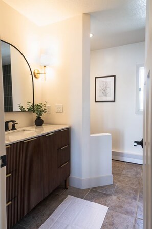 Une salle de bain équipée d’un meuble-lavabo, d’un miroir, d’un luminaire fixé au mur, d’une œuvre d’art encadrée et d’une porte donnant sur une autre pièce.