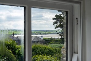 Blick durch ein Fenster auf einen Fluss, Grünflächen und ein Steinhaus.