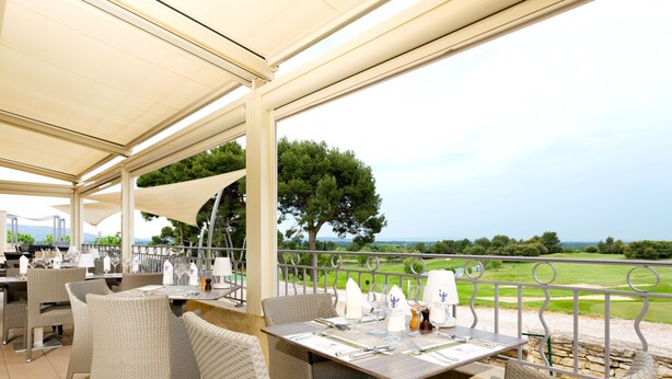 An outdoor dining area with wicker chairs and tables, a covered roof, and a view of a golf course. - Provence Country Club by Interhome (L'isle sur la Sorgue)