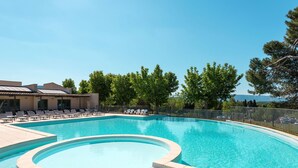 A swimming pool with a curved design, surrounded by lounge chairs and a building with a tiled roof. - Provence Country Club by Interhome (L'isle sur la Sorgue)