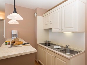 Eine kleine Küche mit weißen Schränken, einem eingebauten Herd und einem Spülbecken. Zwei Pendelleuchten hängen von der Decke, ein Kaffeeautomat steht auf der Arbeitsplatte, und es liegen ein paar Brotscheiben bereit.