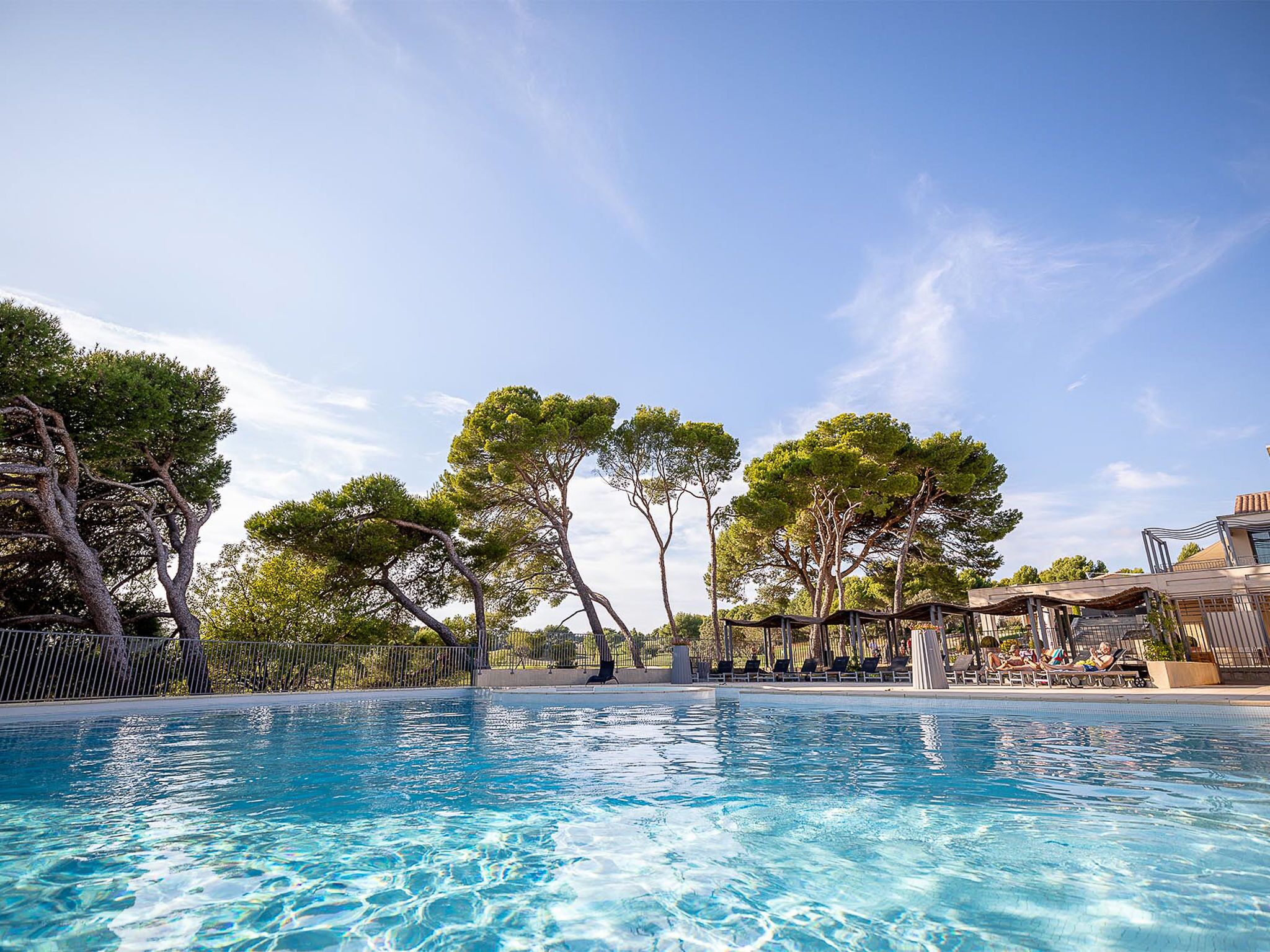 A swimming pool with clear blue water, surrounded by trees and a building with outdoor seating.