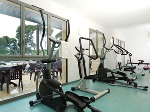 Ein Fitnessraum mit diversen Trainingsgeräten, großen Fenstern und Blick auf eine Außenbestuhlung.