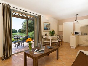 Une cuisine moderne avec des meubles blancs, un coin repas avec une table et des chaises en bois, et une vue sur un jardin grâce à une porte-fenêtre coulissante.