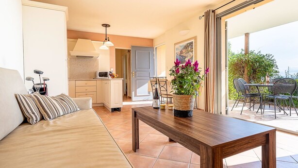 Ein gemütlicher Wohnbereich mit einer Couch, einem Holztisch und einem Esstisch für zwei Personen. Im Hintergrund ist eine Küche zu sehen, und auch ein Balkon mit Sitzgelegenheit ist abgebildet.
