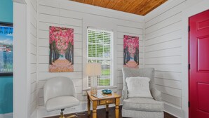 Een kamer met witte lambrisering, een houten plafond, een rode deur, twee stoelen, een klein tafeltje met een lamp en twee ingelijste kunstwerken.