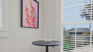 Een kamer met twee houten krukken, een ronde tafel met een glazen vaas, een lijst met een abstract schilderij en een raam met verstelbare zonwering.