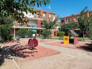A playground with red and yellow equipment, surrounded by a building with balconies and a small red structure. - Proche de la plage | Accès piscine | Parking (ARGELES-SUR-MER)