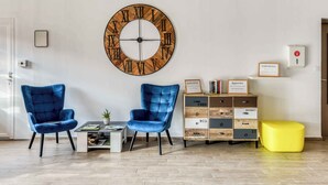 Een woonkamer met twee blauwe stoelen, een salontafel, een grote houten klok, een boekenkast en een gele poef.