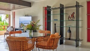 A modern living room with a glass-top dining table, wicker chairs, a flat-screen TV, and a decorative glass shelf with a horse figurine. - Beachfront Resort-Style Villa. Pickleball, tennis, Pools, Restaurant (Cruz de Huanacaxtle)