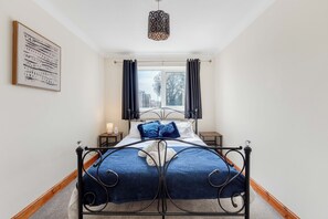 A bedroom with a bed, a window with curtains, a bedside table, and a wall-mounted lamp.