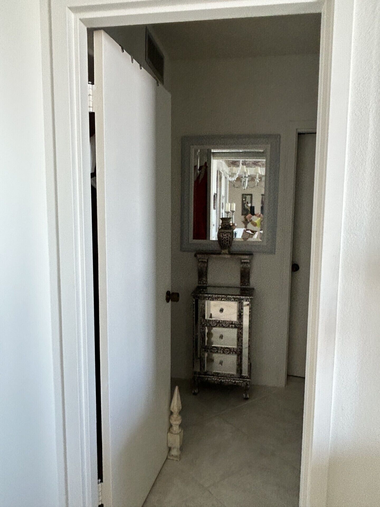 Un couloir avec une porte blanche, un miroir au-dessus d’une commode et un petit objet décoratif posé sur le sol.