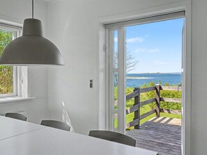 Een kamer met een tafel, stoelen, een hanglamp, een glazen schuifdeur die uitkomt op een terras met uitzicht op zee.